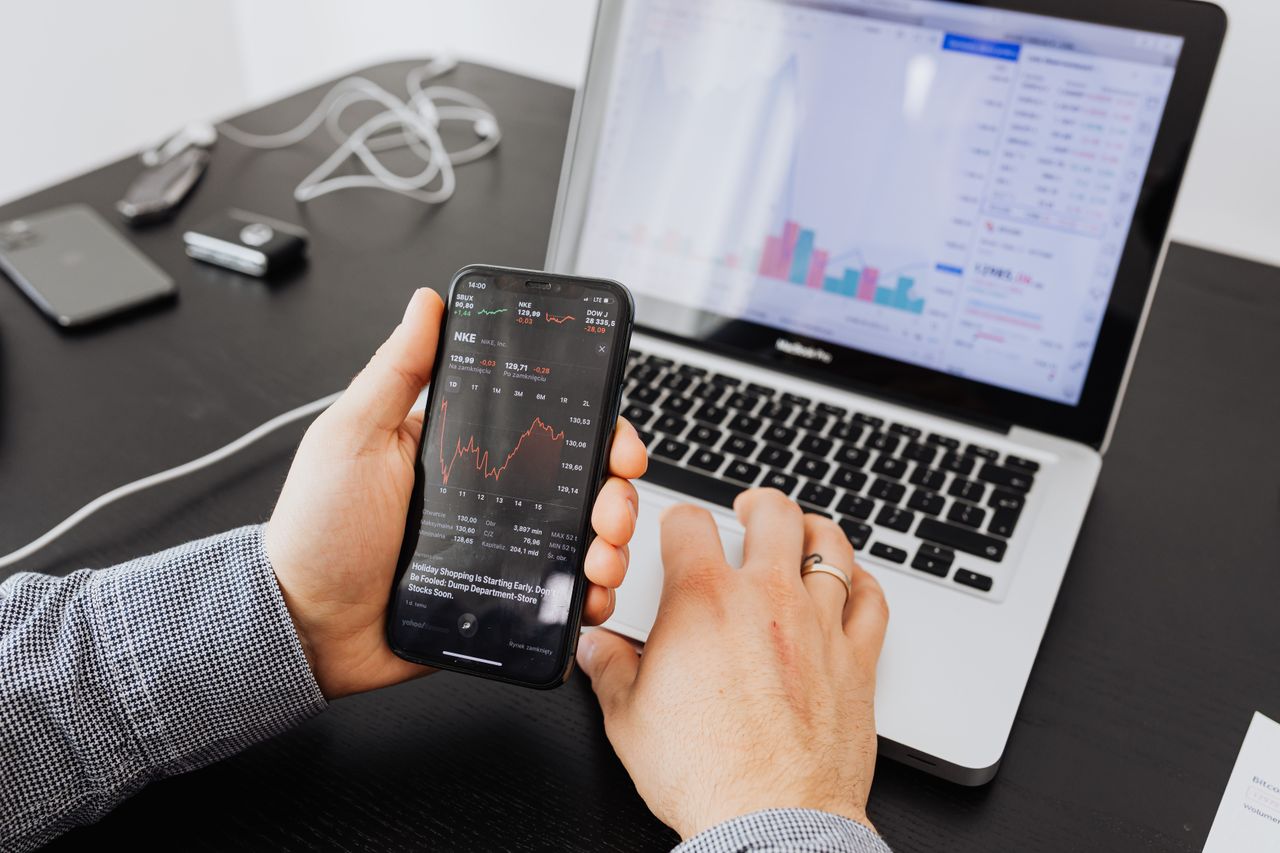 A person studies stock data using a smartphone and laptop indoors.